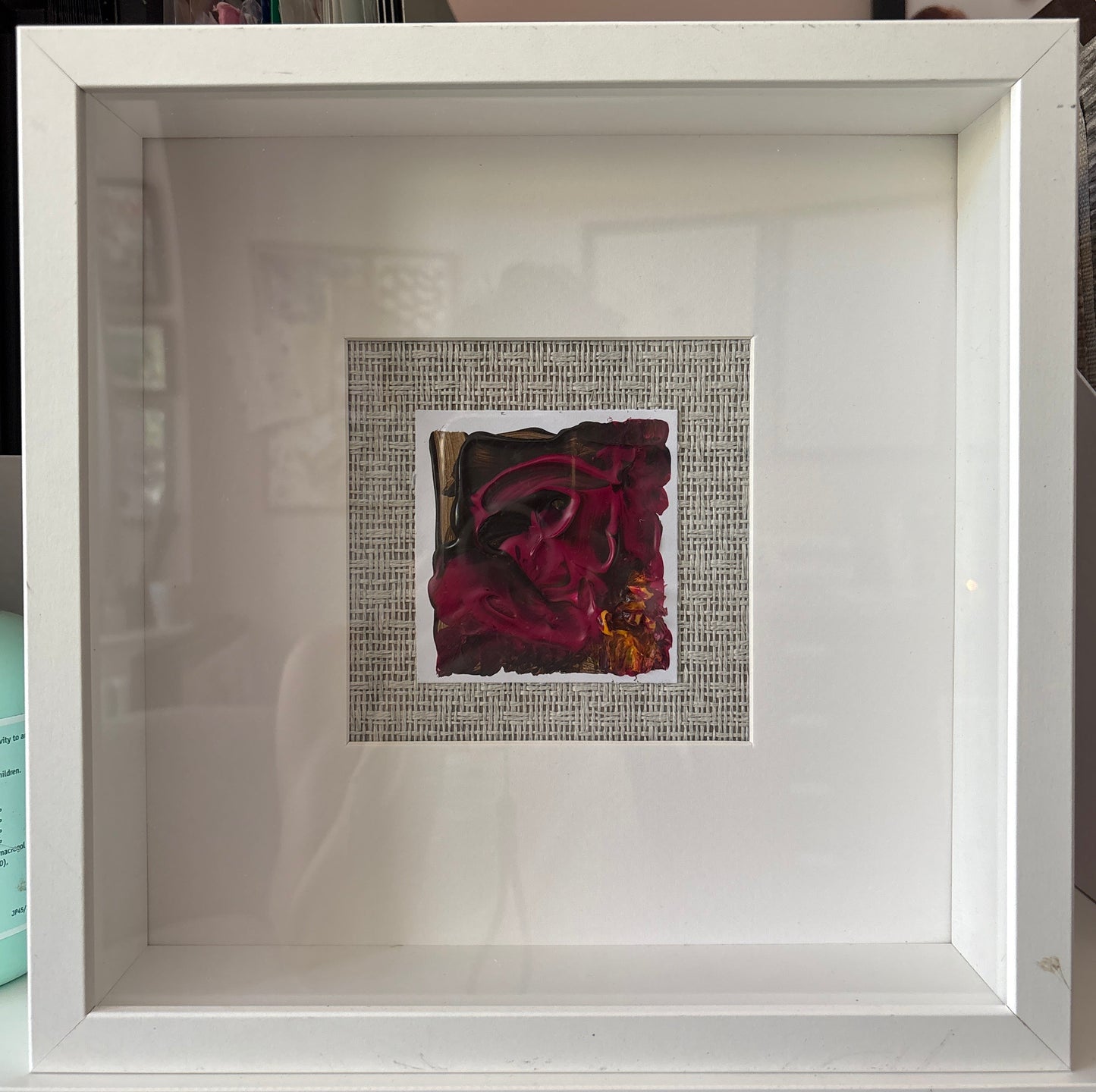 A square white shadowbox frame containing a small central artwork with a dark red abstract floral design mounted on a grasscloth background