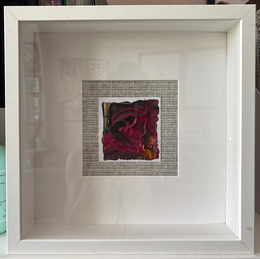 A square white shadowbox frame containing a small central artwork with a dark red abstract floral design mounted on a grasscloth background