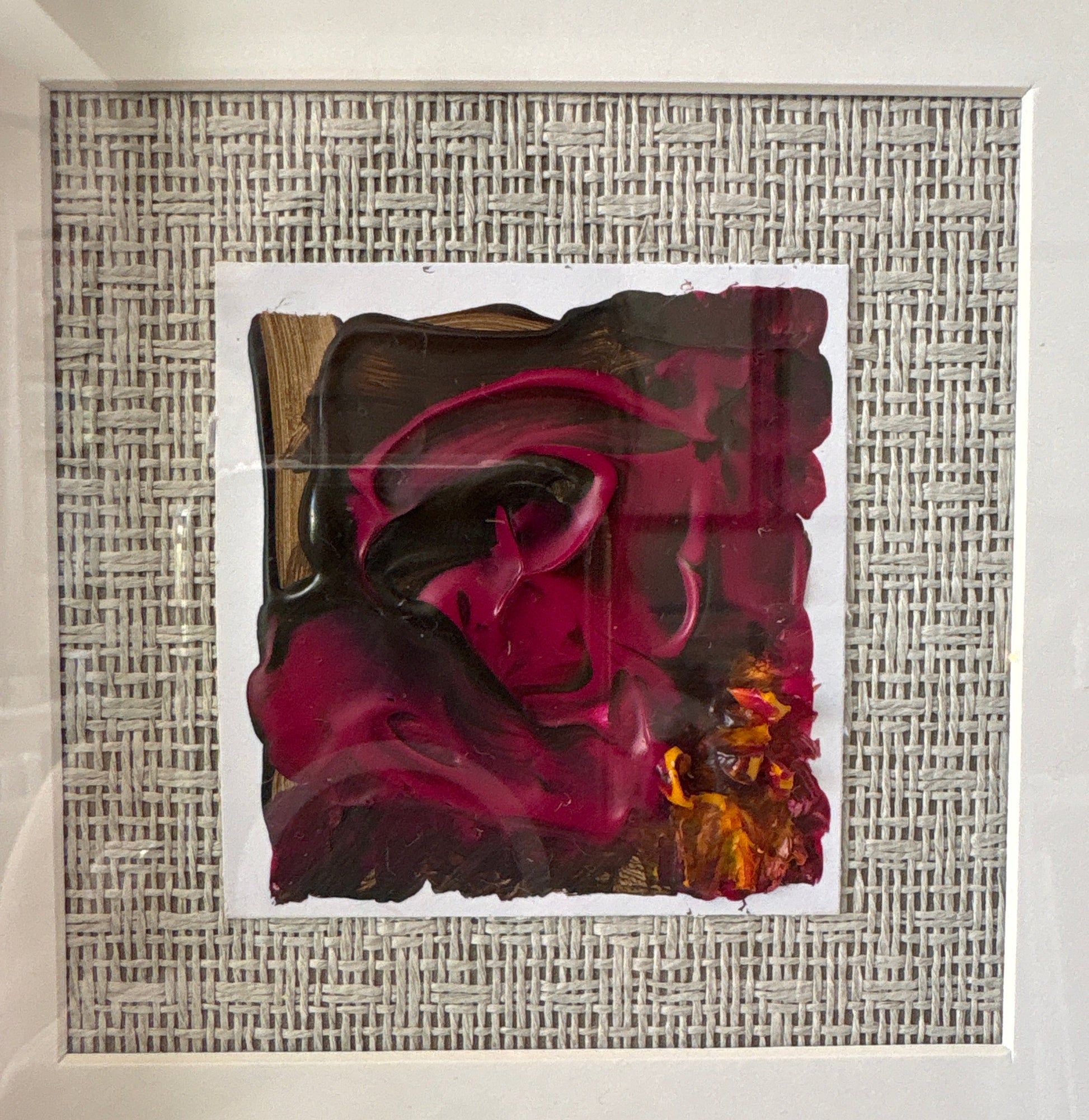A square framed abstract artwork featuring a textured gray and white woven border with a central composition of deep magenta, burgundy, black, and gold paint strokes, enclosed in a clear protective case.