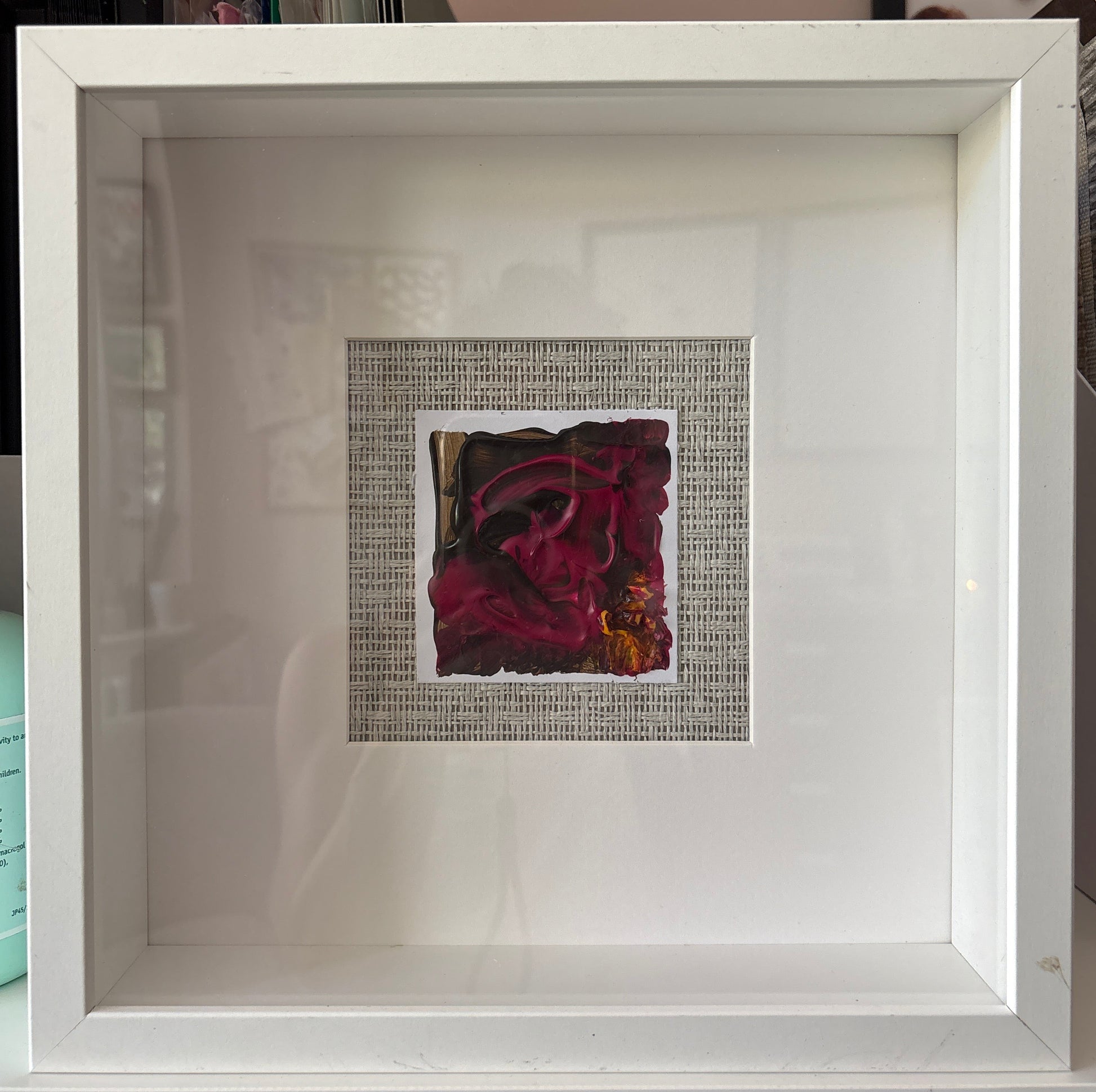 A square white shadowbox frame containing a small central artwork with a dark red abstract floral design mounted on a grasscloth background