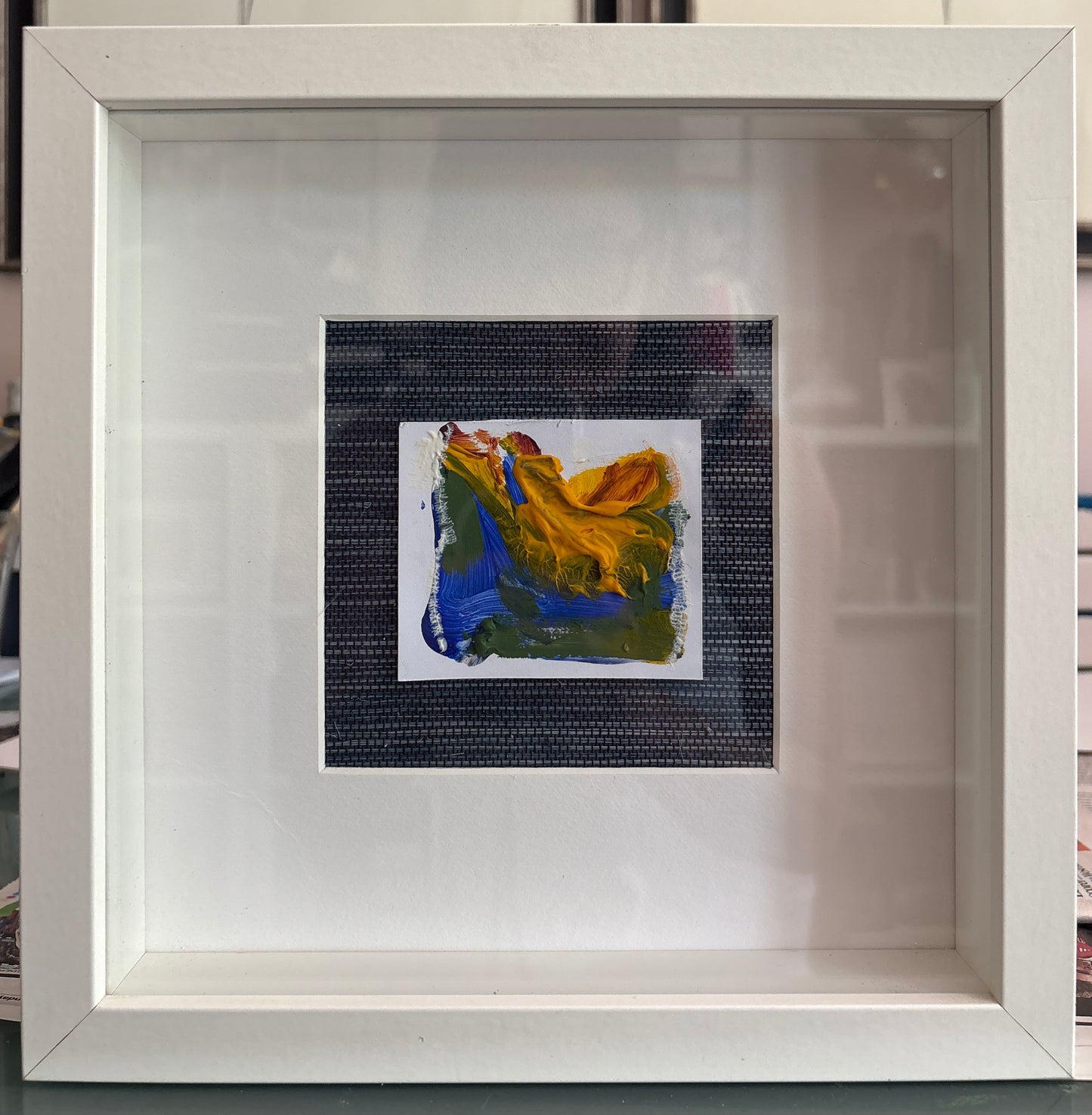 A white square shadow box frame containing a small abstract painting with bold strokes of yellow, blue, green, and white, mounted on a grass cloth background.