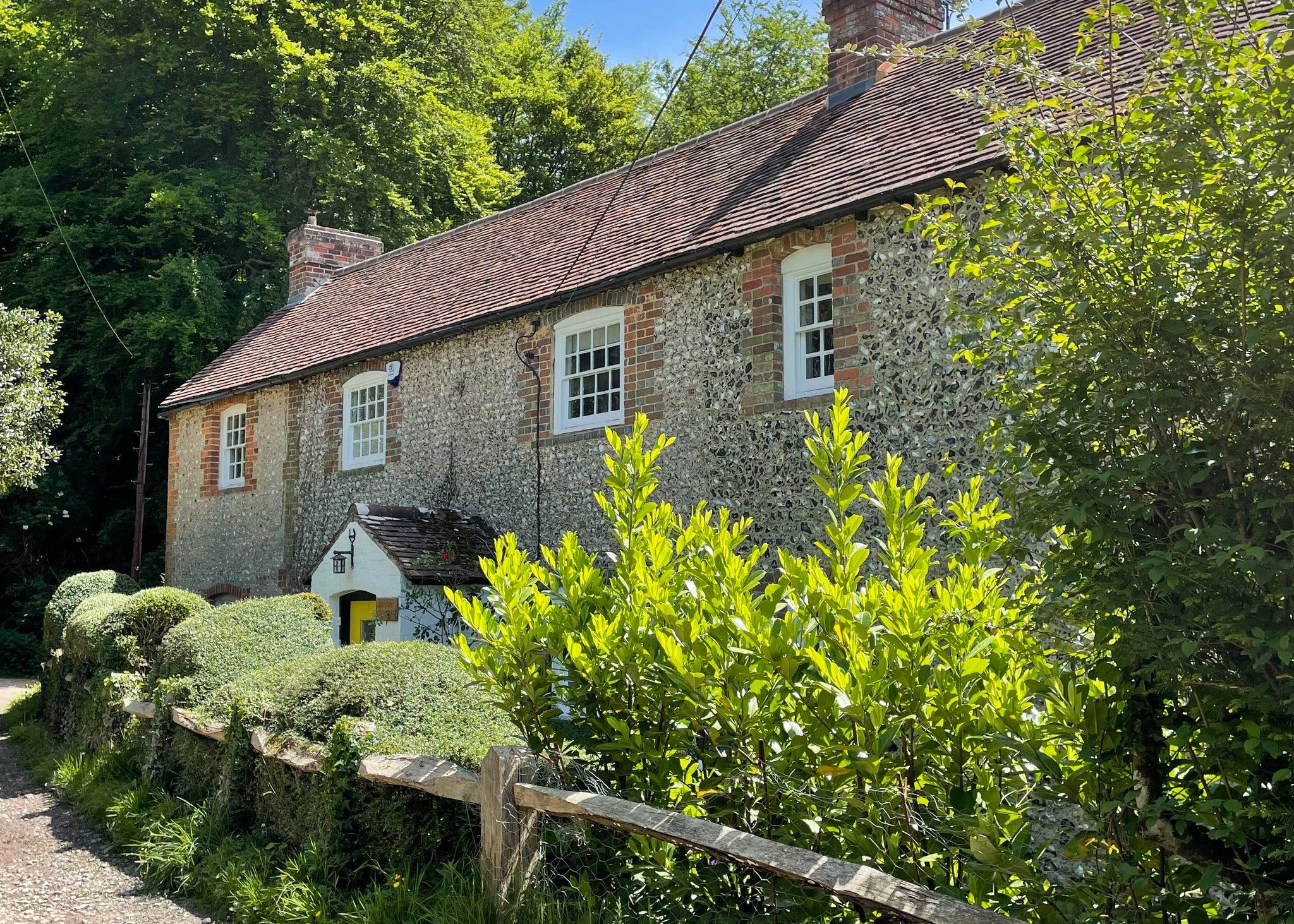 A rustic stone cottage with a brown tiled roof, surrounded by lush green trees and shrubs under a blue sky.